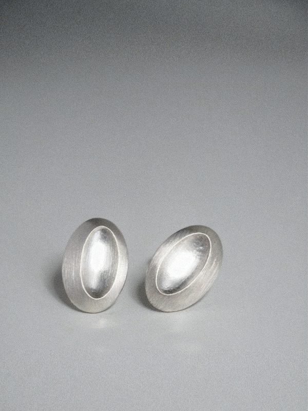 Elongated oval stud earring in polished silver, featuring an off-center tilted 'thumb dent' mimicking the tactile response buttons of vintage MP3 players.