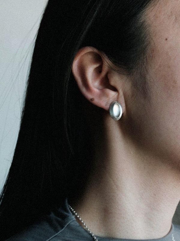 Elongated oval stud earring in polished silver, featuring an off-center tilted 'thumb dent' mimicking the tactile response buttons of vintage MP3 players.