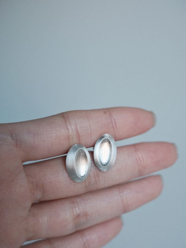 Elongated oval stud earring in polished silver, featuring an off-center tilted 'thumb dent' mimicking the tactile response buttons of vintage MP3 players.